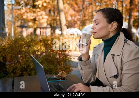 Fröhliche, ruhige, hübsche Frau, die in der Ferne arbeitet, in der hölzernen Cafeteria im Eichenwald chillt und während der Arbeit leckeren heißen Kaffee trinkt Stockfoto