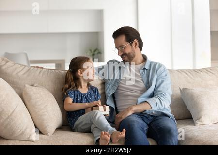 Glückliches kleines Mädchen hält Geschenk, sitzt auf dem Sofa mit Vater. Stockfoto