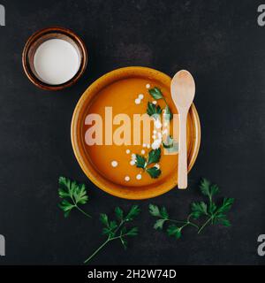 Minimal dunkel flach Lay Idee von hokkaido Kürbiscremesuppe und Petersilienblätter mit Holzlöffel. Heiße Milch oder Joghurt in Keramikbecher. Stockfoto