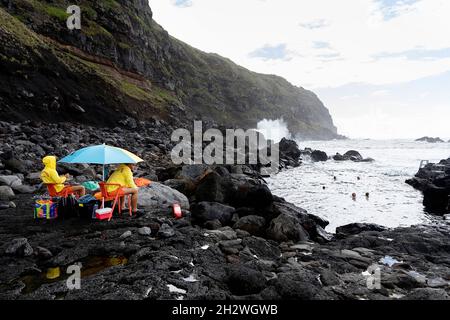 Rettungsschwimmer, die unter einem Regenschirm sitzen und Badenden in vulkanischen Lavaformationen an der Küste von Ferraria, Sao Miguel, Azoren, Portugal, zusehen Stockfoto