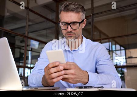 Reifer Geschäftsmann, der ein Mobiltelefon mit dem Telefon hält, um Daten im Büro zu überprüfen. Stockfoto