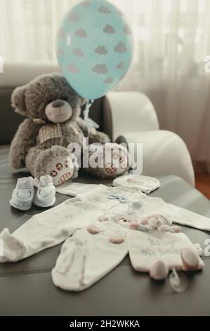 Babyanzüge, Hut, Schuhe und ein Spielzeugbär mit einem blauen Ballon auf dem Bett im Schlafzimmer. Babysachen Stockfoto