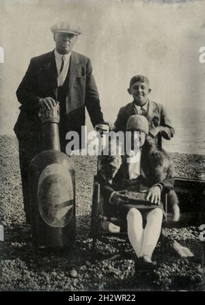 Ein amüsantes englisches „Tintype“-Foto ‘den späten 1920er Jahren, das eine Familie am Strand zeigt. Die Mutter trägt eine Pelzsohle und sitzt auf einem Liegestuhl mit ihrem Sohn hinter ihr. Der Vater hält eine riesige Bierflaschenstütze von ‘Whitbread & Co. London Stout’. Stockfoto