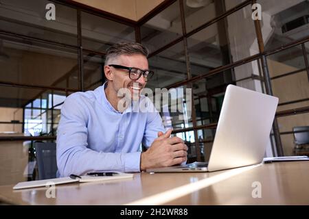 Ein glücklicher, reifer Geschäftsmann, der ein Notebook mit virtuellen Meetings im Büro ansieht. Stockfoto