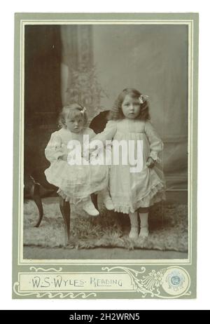 Edwardianische Kabinettkarte Studioportrait von 2 attraktiven entzückenden jungen Mittelklasse-Kindern, Mädchen, die weiße Kleider tragen und die Hände zusammenhalten. Das älteste Mädchen hat ihre Haare gekräuselt und beide tragen Bänder im Haar. Aus dem Fotostudio von W.S. Wyles, Reading, Großbritannien um 1905.