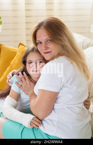 Mutter umarmt Ihr Mädchen mit einem Guss auf einem gebrochenen Handgelenk oder Arm lächelnd. Kinderkrankheiten, positive Aussichten und Genesung. Stockfoto