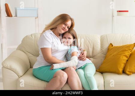 Mutter umarmt Ihr Mädchen mit einem Guss auf einem gebrochenen Handgelenk oder Arm lächelnd. Kinderkrankheiten, positive Aussichten und Genesung. Stockfoto