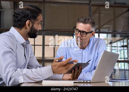 Zwei verschiedene Geschäftsleute diskutieren über Marktdaten mit einem Laptop und einem digitalen Tablet. Stockfoto