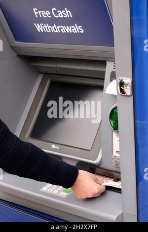 Einer Frau wird gezeigt, wie sie Geld von einem Geldautomaten, einem Geldautomaten oder einem Loch in der Wand nimmt, auf dem ein Schild steht, das anzeigt, dass Geldauszahlungen kostenlos sind. Stockfoto