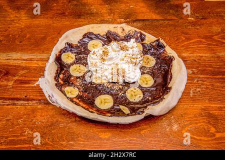 Öffnen Sie auf dem Holztisch Schokoladenkrepe mit Banane, Schlagsahne und Nüssen Stockfoto