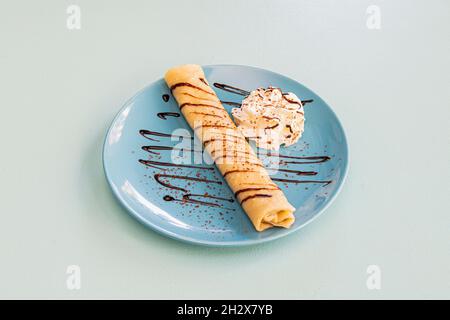 Gerollte süße Crepe, gefüllt mit Schokoladencreme, Schlagsahne, Kakaopulver und Schokoladensirup auf einem blauen Teller Stockfoto