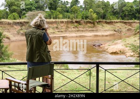 Eine kaukasische Frau steht an einem Geländer und fotografiert Flusspferde am Mara-Fluss in der Ferne, Masai Mara, Kenia Stockfoto