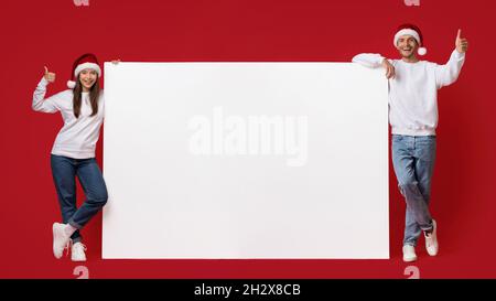 Urlaubsangebot. Fröhliches Paar In Santa Hats, Das In Der Nähe Von Blank Advertisement Board Steht Stockfoto