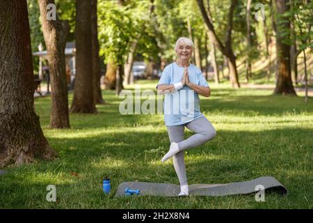 Glückliche ältere Dame im Trainingsanzug tut Baum Pose Yoga Asana im Park zu üben Stockfoto