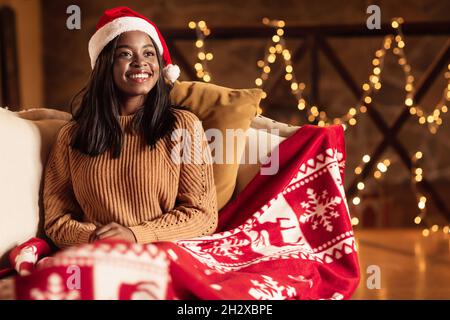 Cozy Christmas atmosphere. Young black woman in Santa hat sitting on couch under warm plaid, celebrating Xmas at home Stockfoto