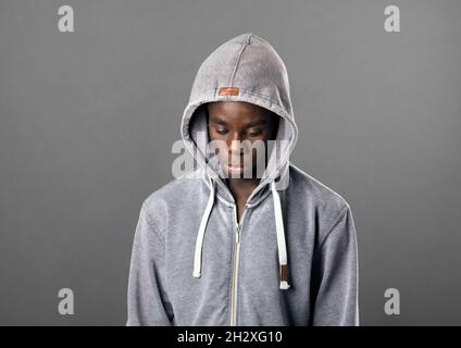 Ein junger schwarzer Mann mit grauem Kapuzenpullover blickt mit einem ernsten düsteren Ausdruck in einem Nahaufnahme-Portrait auf grauem Kopf und Schultern nach unten Stockfoto