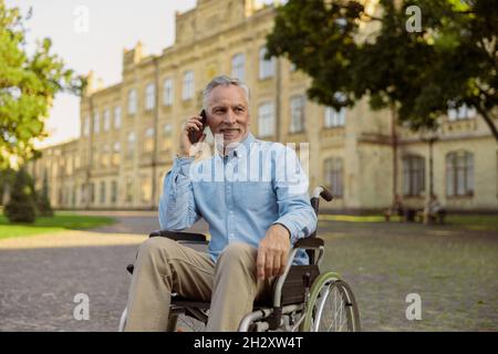 Lächelnder, gealterter Mann, der den Patienten im Rollstuhl wiederfindet und beim Telefonieren wegschaut und Zeit allein im Park in der Nähe des Krankenhauses verbringt Stockfoto