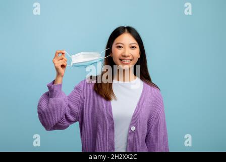 Junge asiatische Frau, die die Einweg-Schutzmaske auszieht, auf blauem Studiohintergrund steht und frische Luft atmet Stockfoto