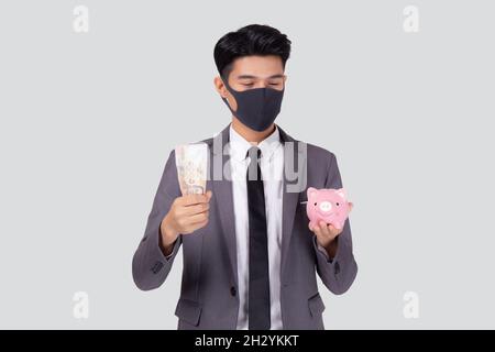 Junge asiatische Geschäftsmann im Gesicht Maske hält Geld Thai und Sparschwein isoliert auf weißem Hintergrund, Krise der Wirtschaft, Geschäftsmann in Maske investme Stockfoto