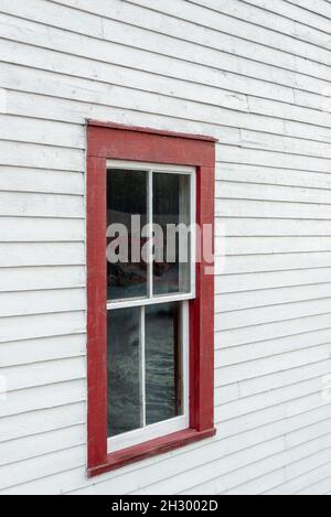Das Äußere einer alten weißen Holzschuppen-Wand mit einem rot umrahmten geschlossenen Glasfenster, das doppelt aufgehängt wurde. Im Fenster ist eine Spiegelung des Himmels. Stockfoto