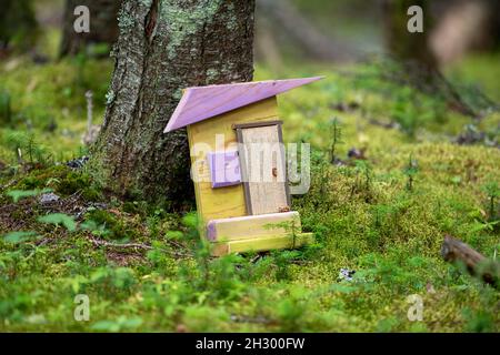 Ein Märchenhaus am Fuße eines Baumes. Es gibt ein kleines gelbes Haus mit einem lila Holzdach, einer einzigen Tür und einem lila Fenster. Stockfoto