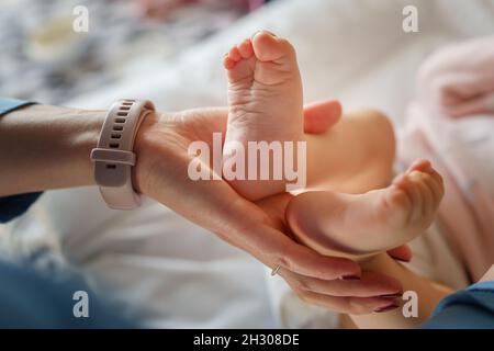 Nahaufnahme der Hände einer unbekannten Frau, die tagsüber die Füße ihres sechs Monate alten Kleinkindes oder ihrer Tochter hält - Mutterschaft und Mutterschaftsliebe Konzept Stockfoto