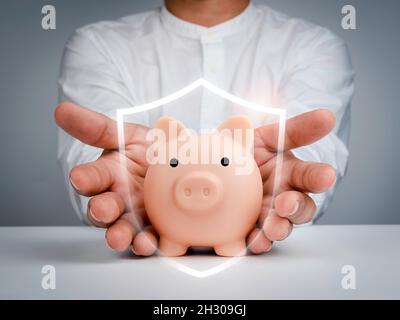 Schutzgeld-Konzept. Das pinke Sparschwein wird durch die Hand eines Geschäftsmannes mit einem Schild-Symbol auf einem weißen Tisch und einem dunkelgrauen Hintergrund geschützt. S Stockfoto