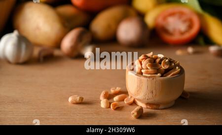 Eine Holzschale aus Erdnüssen auf einem Holztisch mit verschwommenem Gemüse auf dem Hintergrund. Nüsse, Bohnen, gesalzene Erdnüsse. Stockfoto