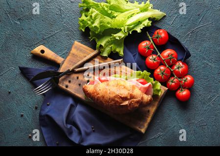Holzbrett mit köstlichem Croissant-Sandwich, Salat und Tomaten auf schwarzem Hintergrund Stockfoto