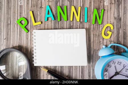 Das Wort Planung besteht aus farbigen Buchstaben, neben einem Notizblock zum Schreiben, einer Lupe und einem Wecker auf einem Holzhintergrund, Geschäftskonzept Stockfoto