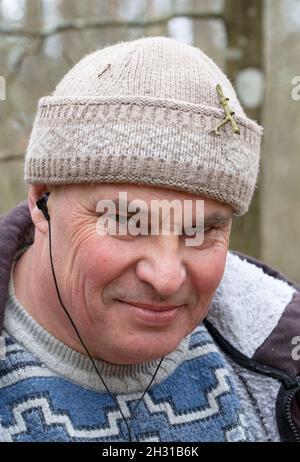 Porträt eines Mannes mittleren Alters in Strickmütze und warmem Pullover, der Musik über Kopfhörer hört und auf die Kamera schaut. Stockfoto