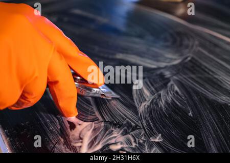 Nahaufnahme eines orangefarbenen Handschuhs mit einem Metallschaber, der das Kochfeld reinigt. Selektiver Fokus. Das Konzept der Hausreinigung eines Elektroofens Stockfoto