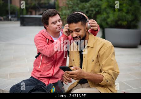 Junger Mann mit Down-Syndrom mit seinem Mentoring-Freund, der im Freien sitzt und Musik hört. Stockfoto