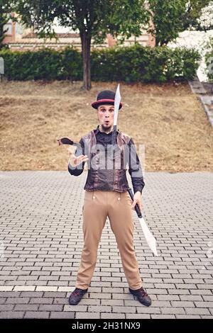 Full Body lustige männliche Darsteller in Kostüm lächelnd und jonglieren Messer, während auf dem Bürgersteig während der Show im Park stehen Stockfoto