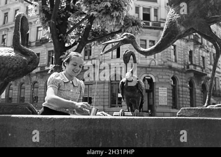 Eine junge Frau probiert das warme Thermalwasser aus dem Reiherbrunnen in der Sophienstraße der Innenstadt von Baden-Baden, Deutschland 1930er Jahre. Eine junge Frau schmeckt das warme Thermalwasser aus der Reiherbrunnen in der Sophienstraße in der Innenstadt von Baden-Baden, Deutschland 1930. Stockfoto