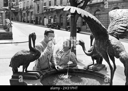 Eine junge Frau probiert das warme Thermalwasser aus dem Reiherbrunnen in der Sophienstraße der Innenstadt von Baden-Baden, Deutschland 1930er Jahre. Eine junge Frau schmeckt das warme Thermalwasser aus der Reiherbrunnen in der Sophienstraße in der Innenstadt von Baden-Baden, Deutschland 1930. Stockfoto