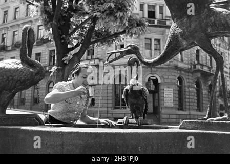 Eine junge Frau probiert das warme Thermalwasser aus dem Reiherbrunnen in der Sophienstraße der Innenstadt von Baden-Baden, Deutschland 1930er Jahre. Eine junge Frau schmeckt das warme Thermalwasser aus der Reiherbrunnen in der Sophienstraße in der Innenstadt von Baden-Baden, Deutschland 1930. Stockfoto