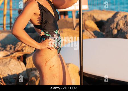 Gesunde Bewegung. Eine gebräunte Frau im Badeanzug steht mit einem Paddel in der Hand vor dem Hintergrund eines Suppboards und zeigt ihre Hüften mit rotem Vess Stockfoto