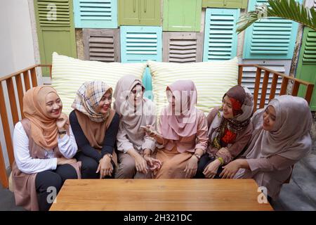 Eine Gruppe asiatischer muslimischer Frauen, die Hijab tragen und auf dem Sofa diskutieren. Smartphone halten und betrachten. Lächelnd und fröhlicher Ausdruck. Stockfoto