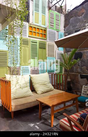 Blick auf das Wohnzimmer im Freien mit gemütlichem Sofa und kopfsteingepflasterter Wand farbenfrohe dekorative Fensterwand. Holztisch und großer Sonnenschirm. Keine Personen. Stockfoto