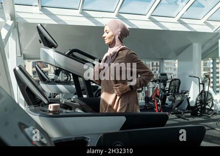 Junge muslimische Frau in Hijab und Sportkleidung, die während des Sporttrainings im Fitnessstudio auf dem Laufband läuft Stockfoto