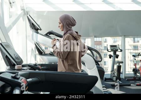 Junge aktive Frau im Hijab und Sportbekleidung, die auf dem Laufband läuft, während sie ihre Freizeit im Fitnessstudio verbringt Stockfoto