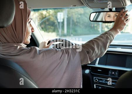 Glückliche junge muslimische Geschäftsfrau in Hijab, die in Spiegel schaut, während sie am Steuer sitzt und das Auto fährt Stockfoto