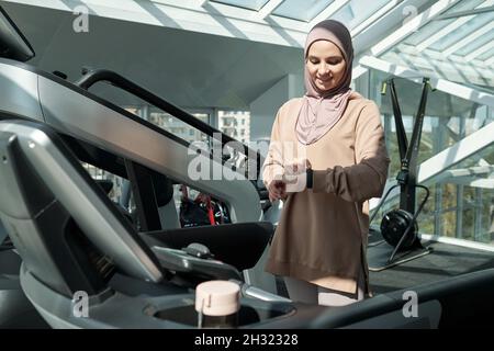 Glückliche junge muslimische Frau, die die Armbanduhr anschaut, während sie am Laufband im großen Fitnesscenter steht Stockfoto