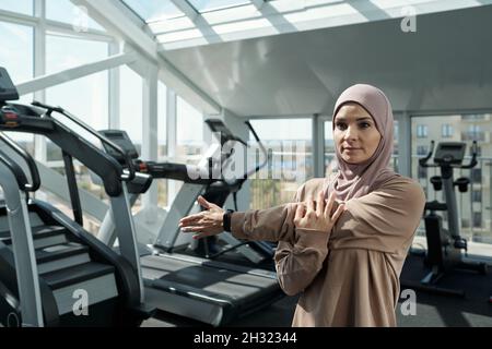 Junge Sportlerin im Hijab, die im Fitnessstudio zwischen Sportgeräten körperliche Übungen für die Arme macht Stockfoto
