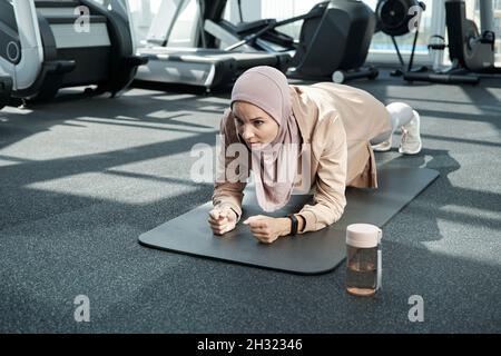 Junge Frauen in Hijab- und Sportkleidung, die sich während der Plank auf der Matte im Fitnesscenter oder Fitnessstudio anstrengen Stockfoto