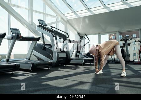 Junge muslimische Frau in Hijab und Sportkleidung, die sich während des körperlichen Trainings im Fitnesscenter nach vorne beugt Stockfoto