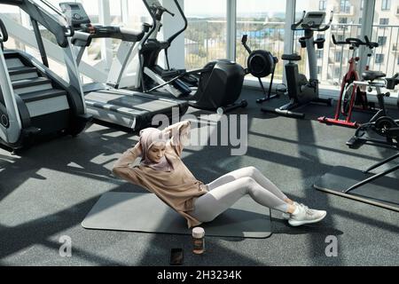 Junge, hübsche Sportlerin, die Sit-Ups mit Seitenwechseln macht, während sie auf der Matte sitzt und ihre Hände hinter dem Kopf hält Stockfoto