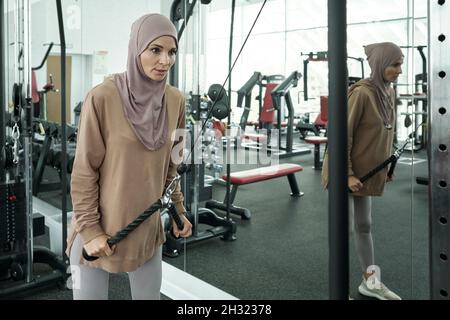 Junge, starke Sportlerin, die am Griff der Sportgeräte zieht, während sie im Fitnessstudio am großen Spiegel steht Stockfoto