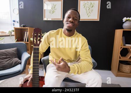 Fröhlicher junger afrikanischer Musiklehrer mit Gitarre, die vor der Kamera gegen das Innere des Hauses sitzt Stockfoto